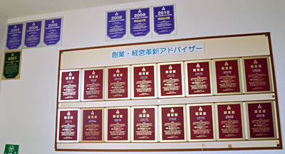 所内に掲示されている「創業・経営革新アドバイザー認定証」や「翌月巡回監査率90%超達成証」のパネル